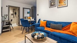 Una sala de estar moderna con un sofá azul, cojines naranjas, una mesa de centro con objetos decorativos y un comedor con sillas azules.
