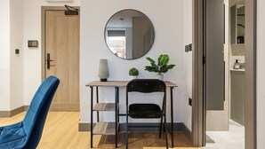 Una habitación moderna con puerta de madera, un espejo redondo en la pared, una mesa pequeña con una planta, una silla azul y vista a otra habitación.