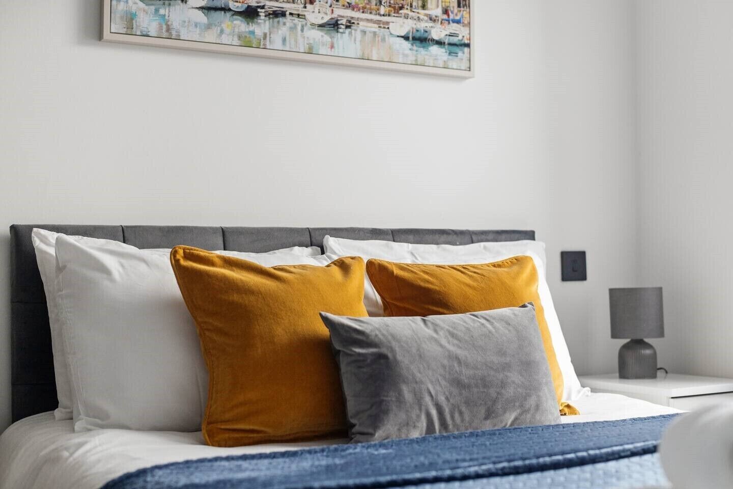Een net opgemaakt bed met witte en mosterdkleurige kussens, een grijs hoofdbord en een blauwe deken.