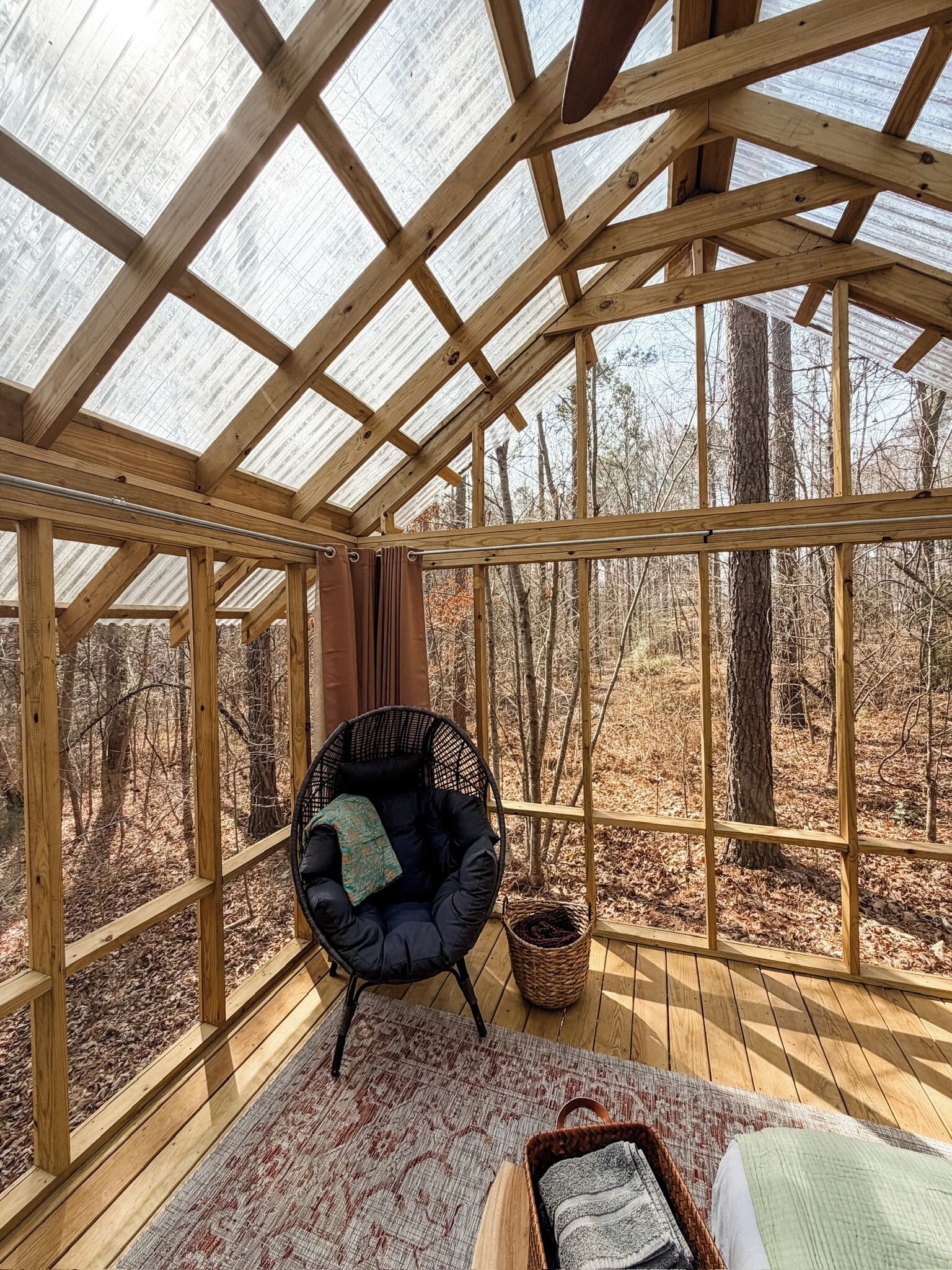 Eine Holzterrasse mit Glasdach, ein schwarzer Stuhl, ein gewebter Korb und ein Bett mit Decke.