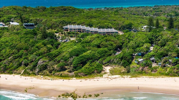 An aerial view of a resort with a large building surrounded by dense forest, a sandy beach, and the ocean. - Welcome to James Cook 2. A beautifully renovated, light-filled townhouse in an unbeatable location. (Byron Bay)