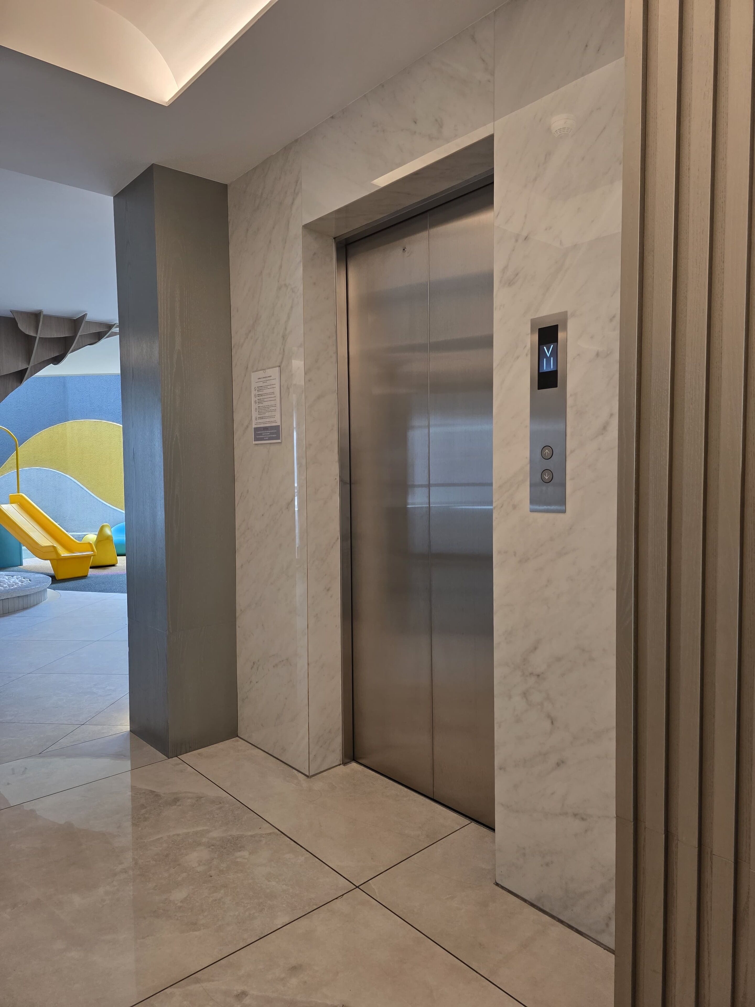 Un ascenseur moderne doté d’un panneau de commande, de murs en marbre et d’un espace de jeux avec des équipements jaunes.