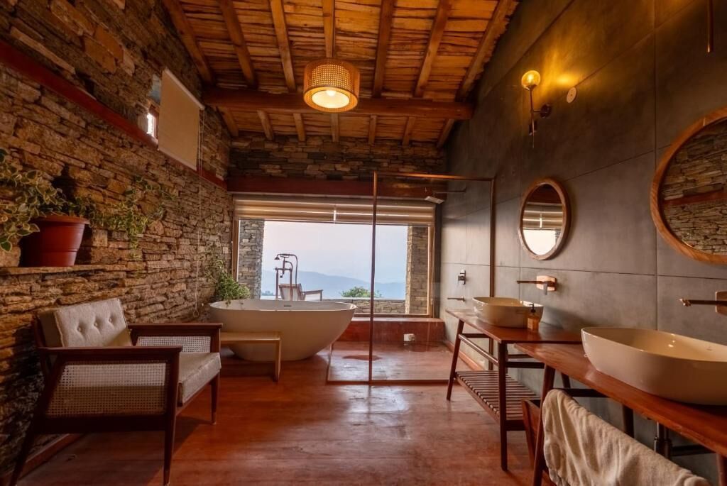 Bagno con parete in pietra, soffitto in legno, una vasca da bagno ampia, un mobile bagno in legno e vista attraverso una porta di vetro.