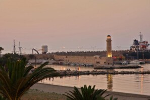 A coastal scene with a prominent lighthouse, a fortified wall, and a calm harbor with boats. - Nonna Irene | ILARIA by CVR (Rethymno)