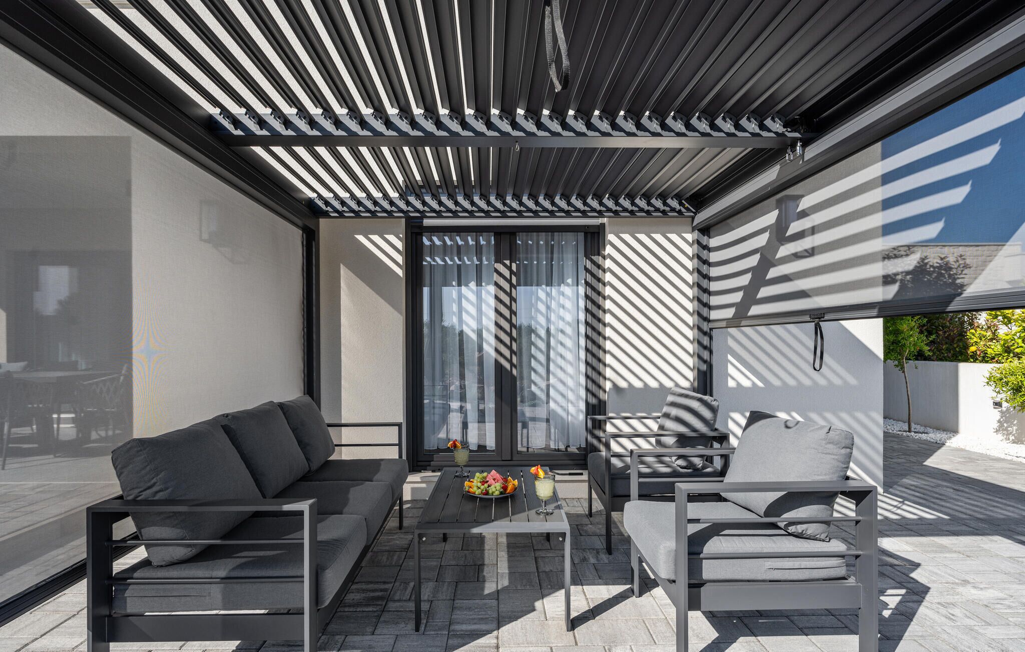 A modern outdoor seating area with a sofa, chairs, and a table, featuring a pergola overhead and large windows.