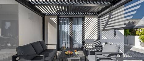 A modern outdoor seating area with a sofa, chairs, and a table, featuring a pergola overhead and large windows.