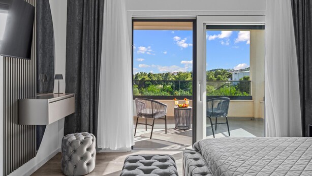 Una moderna camera d'hotel con un letto, una TV, un balcone con sedie e un tavolo e vista sul verde.