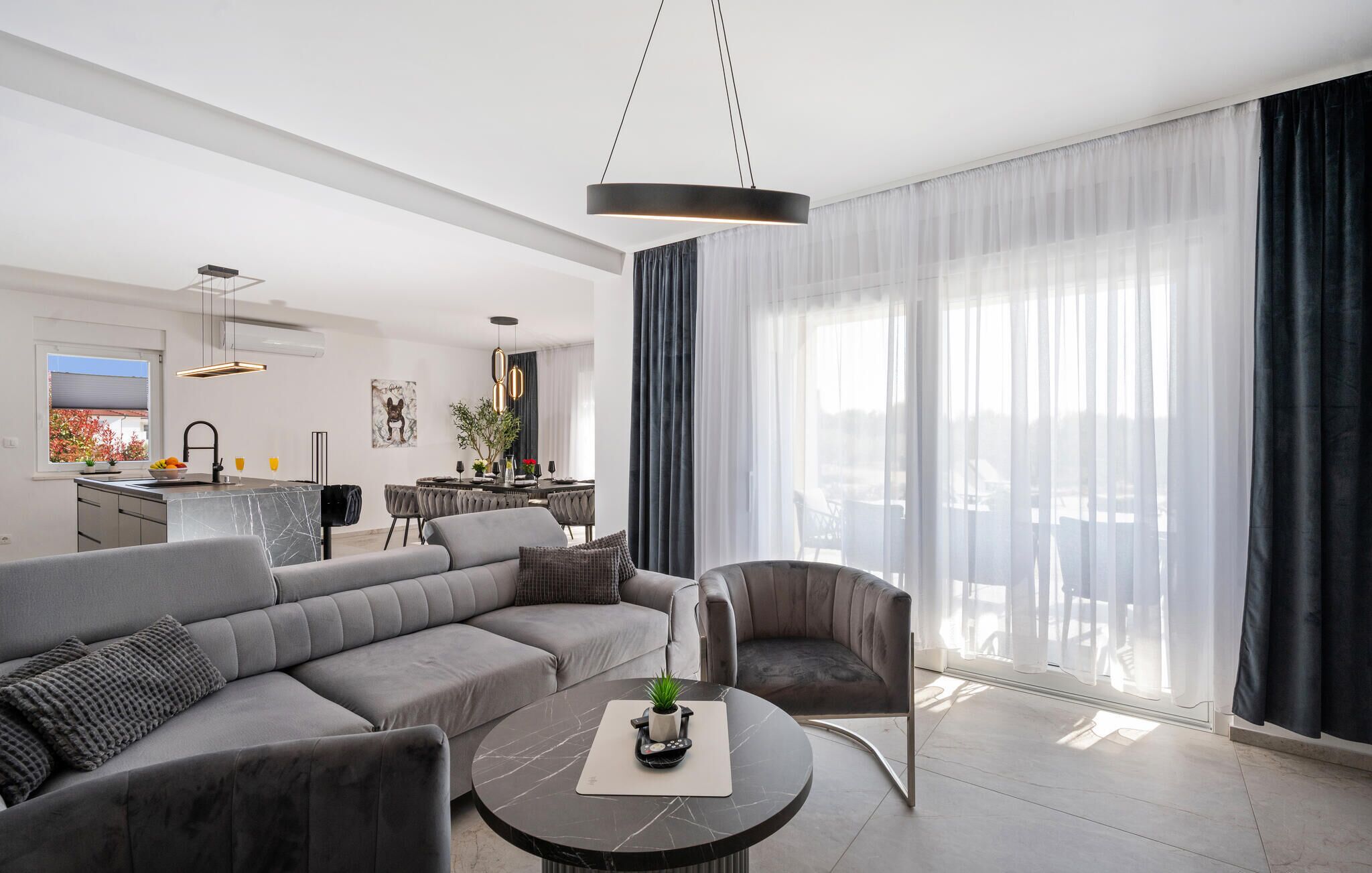 A modern living room with a grey sofa, a round coffee table, and a dining area with a kitchen in the background.