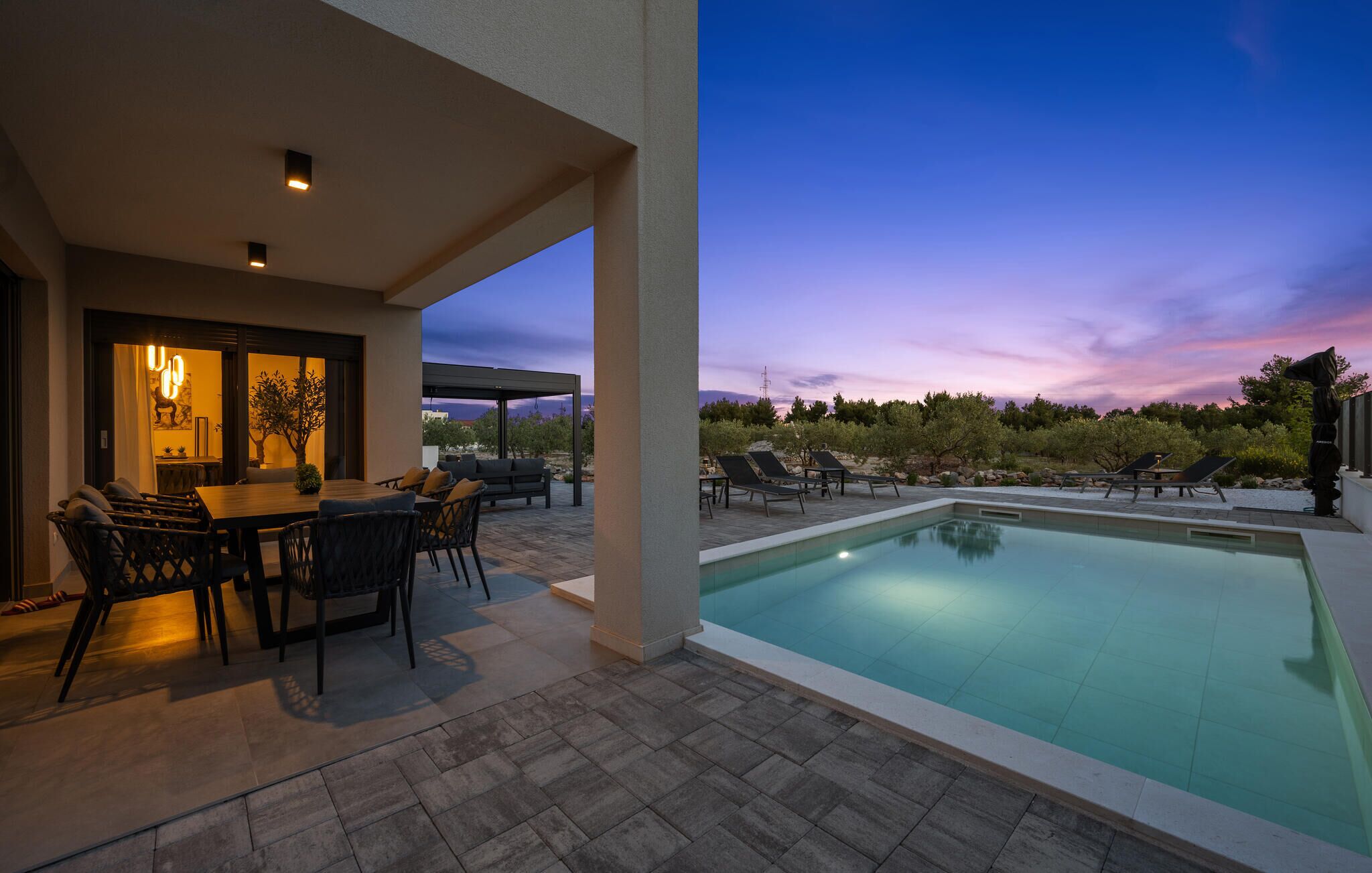 A pool area with a table and chairs, a covered seating area, and a view of the sky.