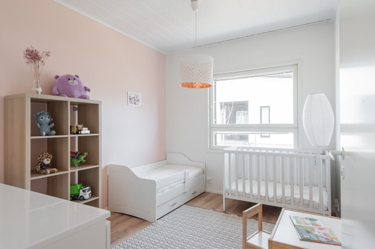 A child's room with a crib, a white bookshelf with toys, a small white bed, and a window with a view.