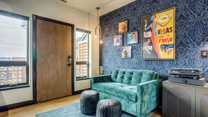 Una habitación con un sofá azul turquesa, dos otomanas, un papel tapiz estampado, una puerta y un mueble con un altavoz.