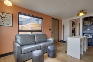 A modern living room with a grey leather sofa, a small ottoman, a white table, and a large window with a view of buildings. - The Louie Boutique Motel (Newport)