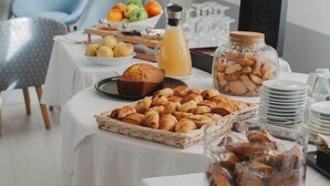 Ein Frühstücksbuffet mit verschiedenen Backwaren, Früchten und Getränken auf einem Tisch in einem gut beleuchteten Raum, im Hintergrund ein Stuhl und ein Schreibtisch.