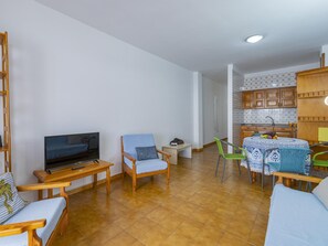 A living room with a TV, wooden furniture, a dining area with a table and chairs, and a kitchen with wooden cabinets.