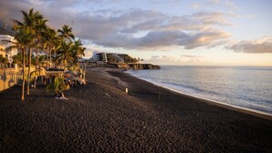 A beach with palm trees, a building with balconies, and a view of the ocean. - CHL El Faro, next to the Puerto Naos beach (Puerto Naos, Los Llanos de Aridane)