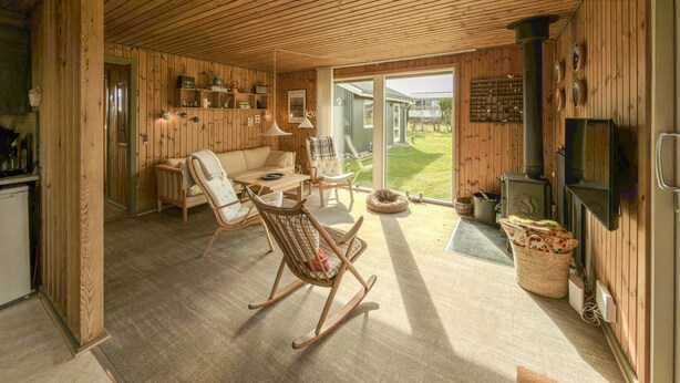 Ein gemütliches Blockhaus-Interieur mit Holzausstattung, einer Couch, Sesseln, einem Esstisch, einem Kamin und Blick ins Freie.
