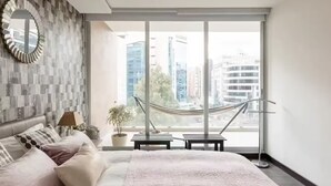 Un dormitorio moderno con una cama grande, vistas de la ciudad a través de ventanales del suelo al techo, una hamaca y una planta en maceta.