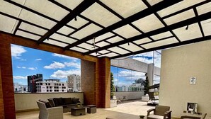 Un patio al aire libre con mobiliario de mimbre, techo con diseño de cuadrícula y vista a edificios al fondo.