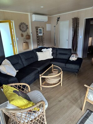 Ein Wohnzimmer mit einem dunkelgrauen Ecksofa, einem Korbstuhl, einem Couchtisch mit Glasplatte und einer Tür, die in einen anderen Raum führt.