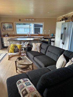 Ein modernes Wohnzimmer mit einem dunkelgrauen Ecksofa, einem Glastisch und einem Korbstuhl. Im Hintergrund ist eine Küche mit einem Edelstahlkühlschrank und einem Fenster zu sehen.