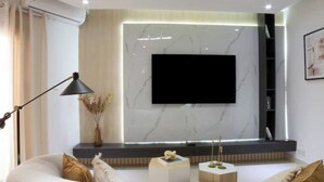 A modern living room with a flat-screen TV mounted on a marble wall, a sofa with cushions, a floor lamp, and a small wooden table with decorative items. - Luxurious 1-bedroom apartment & 1 African-style bedroom (Dakar)