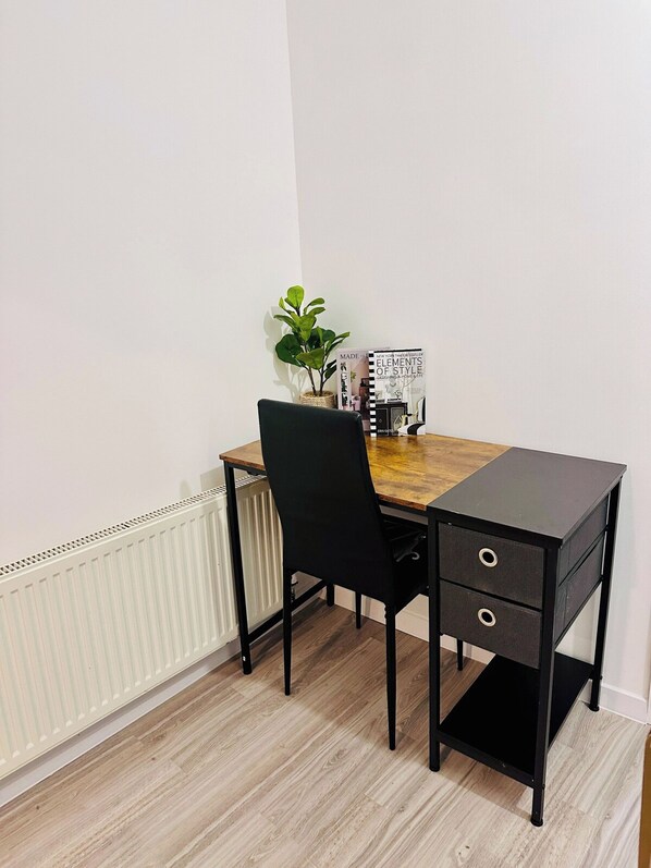 A small, neatly arranged study area with a wooden desk, a black chair, a radiator, and a potted plant.