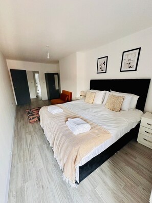 A modern bedroom with a large bed, a nightstand, a chair, and a door leading to another room.