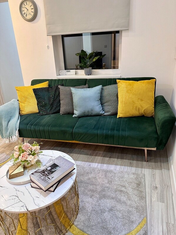 A green sofa with multiple cushions, a round coffee table with a book and flowers, and a clock on the wall.