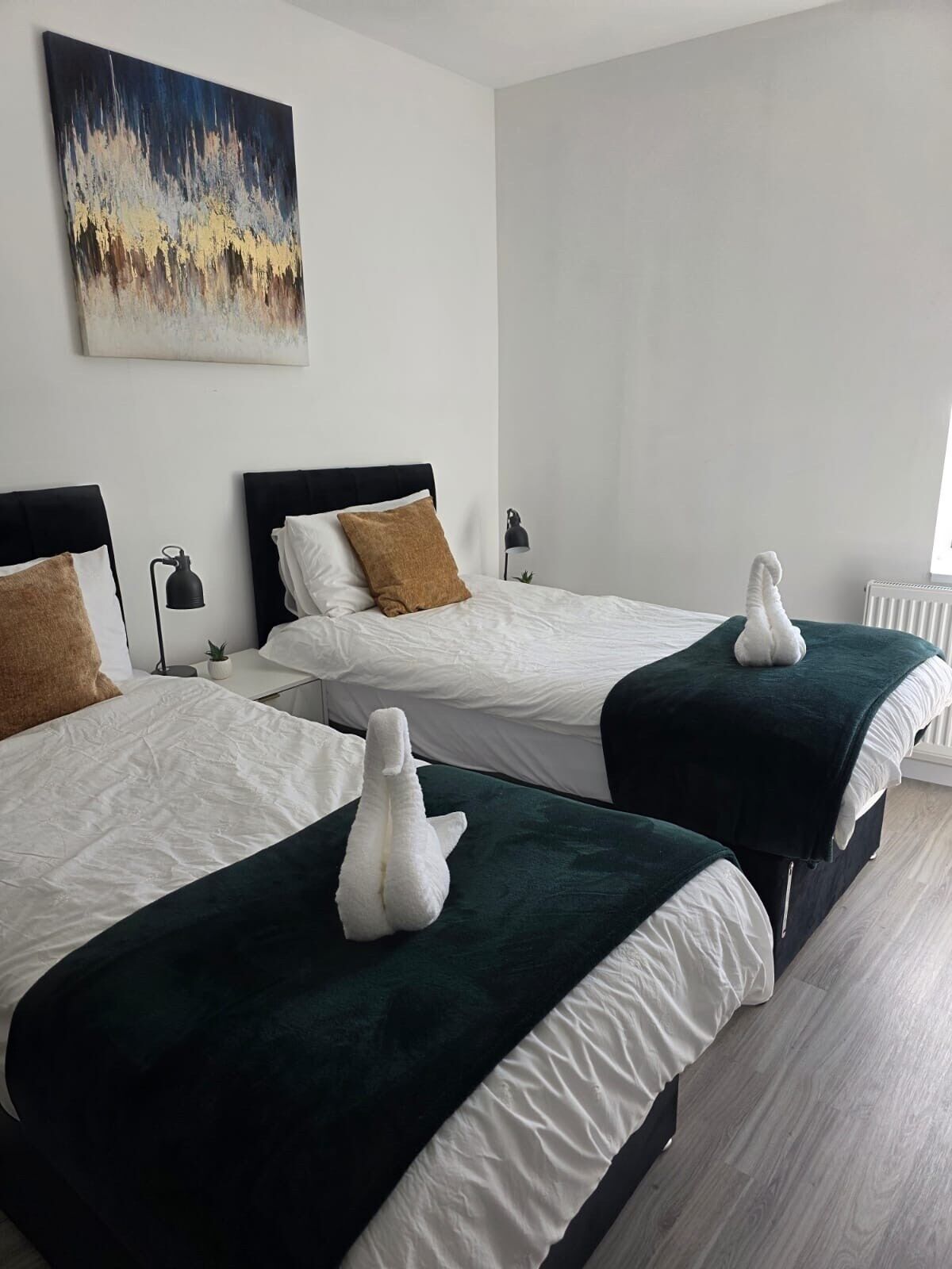 Ein Hotelzimmer mit zwei Betten, jeweils mit weißer Daunendecke und dunkelgrüner Tagesdecke. An der Wand über den Betten hängt ein Bild, auf dem Nachttisch steht ein kleiner Topfpflanze und auf dem Bett liegt ein weißes Handtuch.