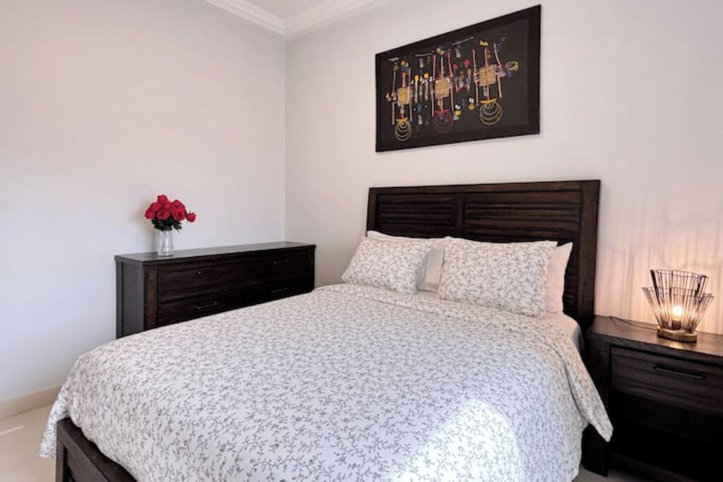 Un dormitorio con cama, mesita de noche, cómoda, un jarrón con flores rojas y una obra de arte abstracto colgada en la pared.