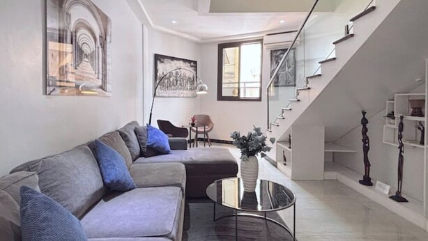 Ein modernes Wohnzimmer mit einer grauen Eckcouch, einer Glaskonstruktion als Treppe und Kunstwerken an den Wänden.