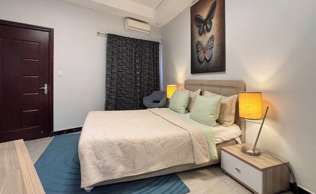 Quarto com cama, luminárias de cabeceira, uma porta, janela com cortinas e uma obra de arte na parede.