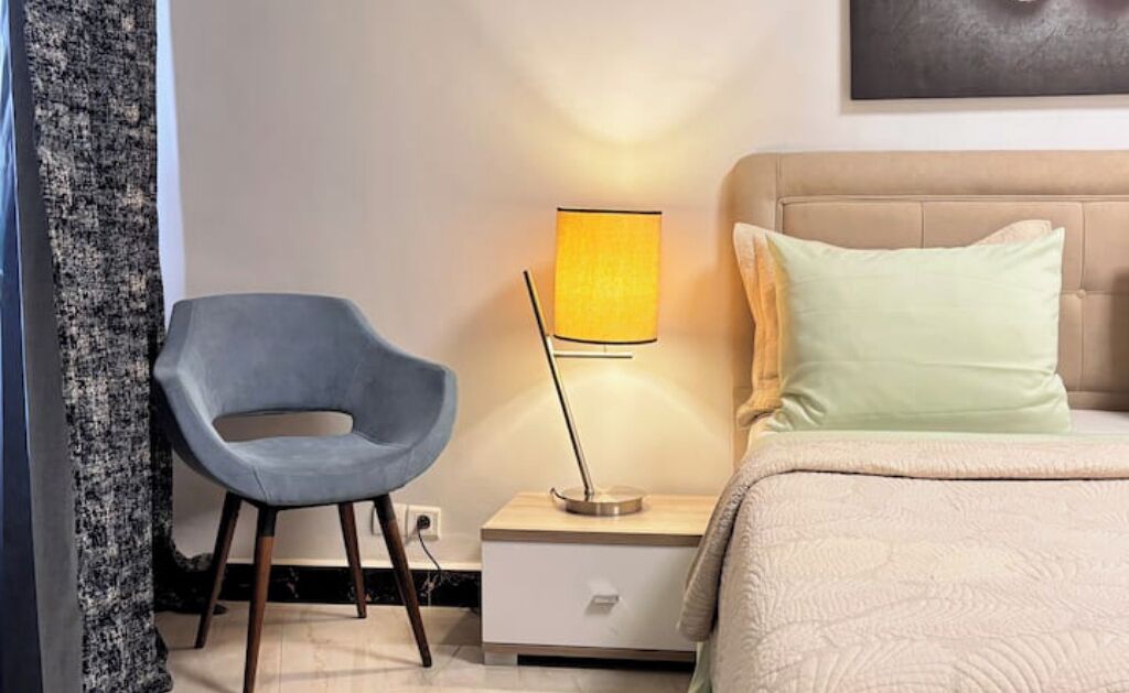 A bedroom with a bed, a nightstand, a lamp, a chair, and a wall-mounted artwork.
