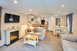 A modern living room with a fireplace, a flat-screen TV mounted above it, a grey sofa, a wooden coffee table, and a dining area with a white table and chairs. - Host Stay Serenity Lodge (York)