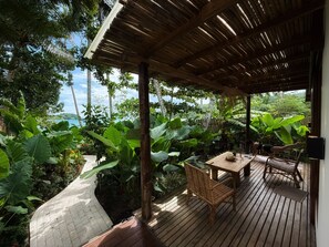Eine Holzterrasse mit Tisch und Stühlen, umgeben von üppiger tropischer Vegetation und Blick aufs Meer.