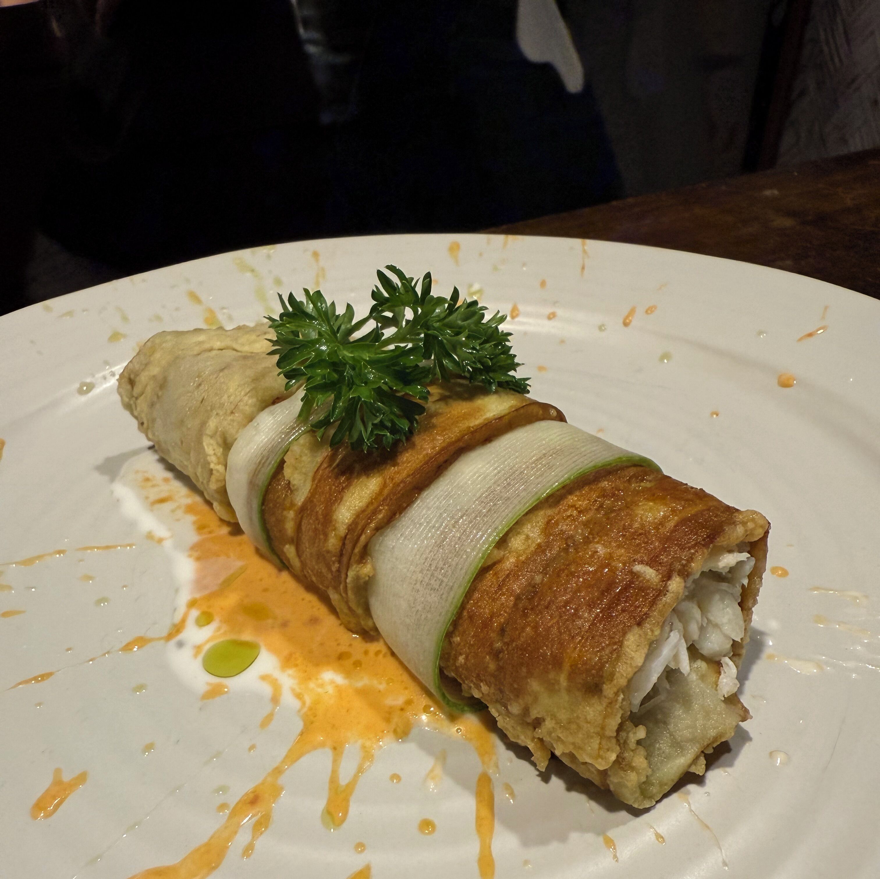 A plate with a rolled dish, possibly a pastry, garnished with green herbs and drizzled with a yellow sauce.