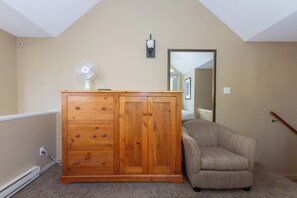 Ein Zimmer mit einem Holzschrank, einem beigen Sessel, einem Standventilator und einem Spiegel an der Wand.