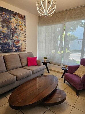 A modern living room with a grey sofa, a round wooden coffee table, a purple armchair, a small side table with a plant, and a large abstract painting on the wall. - Vacation home 5 minutes from the beach, ideal for a family. (Mezcales)