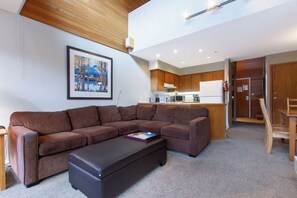 A modern living room with a brown sectional sofa, a black ottoman, and a framed painting on the wall. - Spacious Whistler Village Condo w/ Private Hot Tub | Prime Location Year-Round (Whistler)