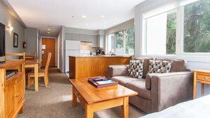 A modern living room with a sofa, coffee table, and wooden furniture. A kitchen is visible in the background with a large window. - Whistler Escape w/ Slope Access | Walk to Lifts & Village Fun | Near Gondola (Whistler)