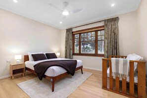 A bedroom with a bed, bedside tables, a crib, a window with curtains, and a ceiling fan.