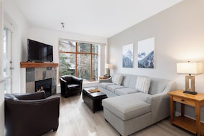 Ein modernes Wohnzimmer mit Kamin, Fernseher, einem grauen Sofa, einem schwarzen Ledersessel und einem Holztisch.