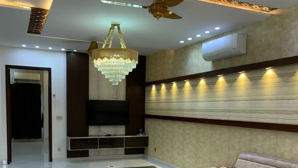 Een moderne woonkamer met een beige bank, een kroonluchter, inbouwspots, een plafondventilator en een flatscreen televisie aan de wand bevestigd.