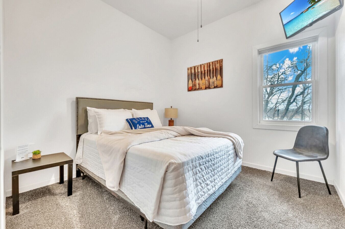 A bedroom with a bed, a chair, a nightstand, a window with a view, and a painting on the wall.