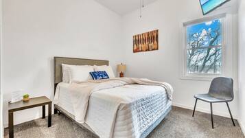 A bedroom with a bed, a chair, a nightstand, a window with a view, and a painting on the wall.