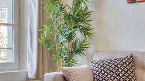 Ein Wohnzimmer mit einem beigefarbenen Sofa, einem gemusterten Kissen, einer Bambuspflanze und einem Fenster mit Vorhängen.