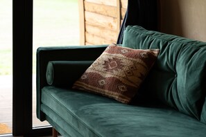 A green sofa with a patterned pillow, positioned near a window with a view of a wooden structure outside. - Luxury Rural Cabin with Wood Fired Hot Tub (Driffield)