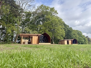 A rustic wooden cabin with a large arched window and a chimney, surrounded by a grassy field and dense trees.
