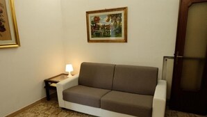 Ein Zimmer mit einer Couch, einem kleinen Tisch mit einer Lampe, einem gerahmten Bild an der Wand und einer Tür.