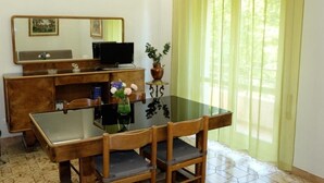 Ein Zimmer mit einem Holztisch, einem Computer, einem Glastisch und Stühlen. Ein Fenster mit Vorhängen ist vorhanden.
